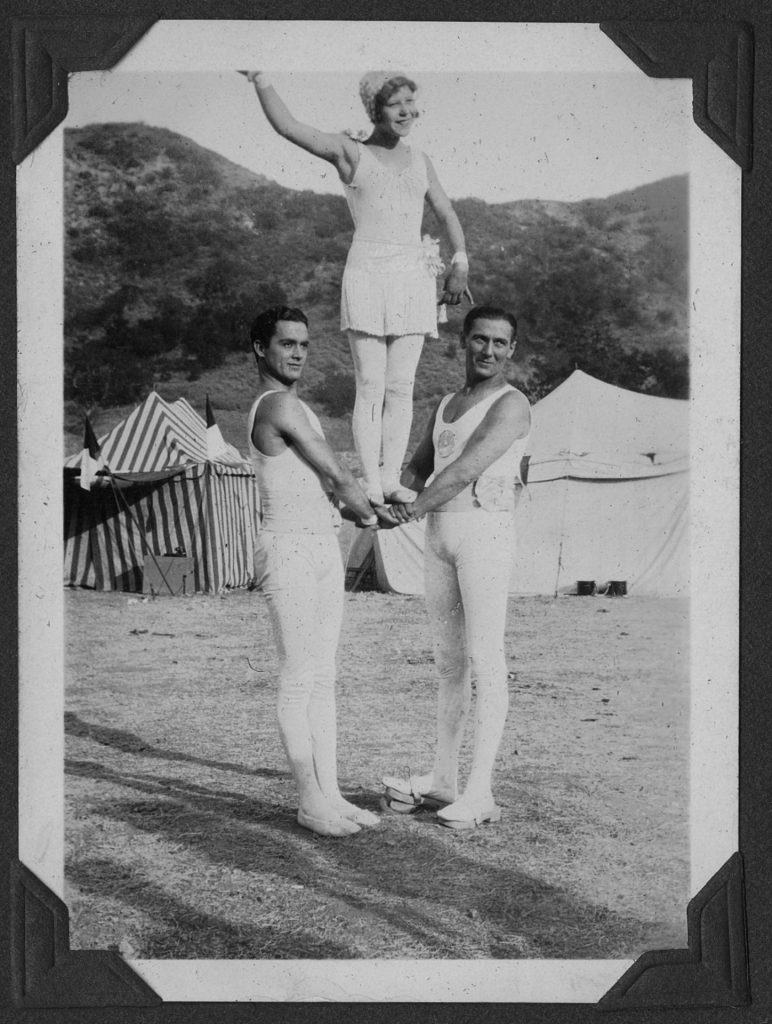 A young woman in a short white dress and spangled cap stands on the joined hands of two men. Circus tents can be seen in the background.