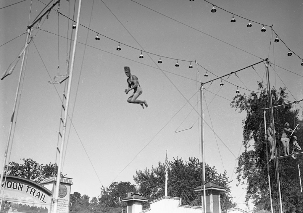 A man in a sparkly leotard is suspended in mid-air between two trapeze swings. 