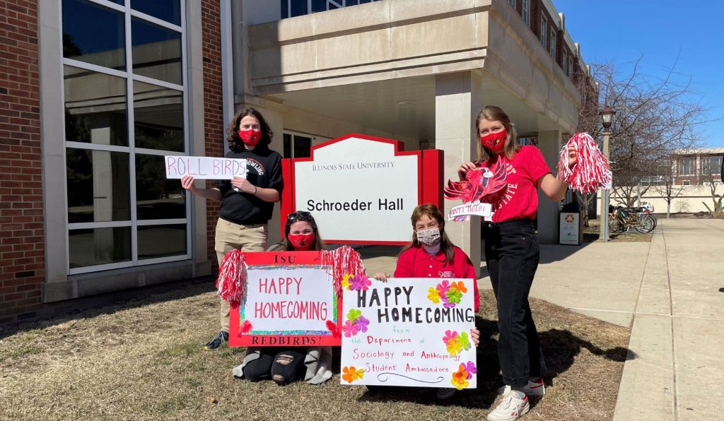 Four SOA student ambassadors with Homecoming signs in front of Schroeder Hall.