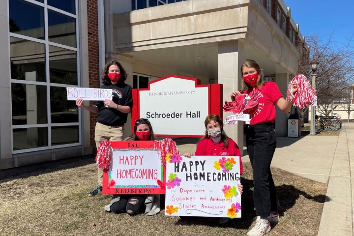 Four SOA student ambassadors with Homecoming signs in front of Schroeder Hall.