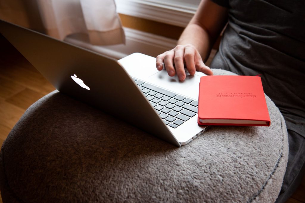 An opened Apple MacBook on a cushioned table with a red notebook balancing on the edge of the keyboard and an individual using the trackpad with their fingers