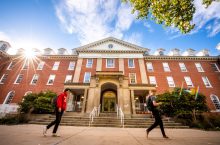 Two male students walk from left to right on Illinois State University's Quad in front of Fell Hall, a brick building below a blue sky.