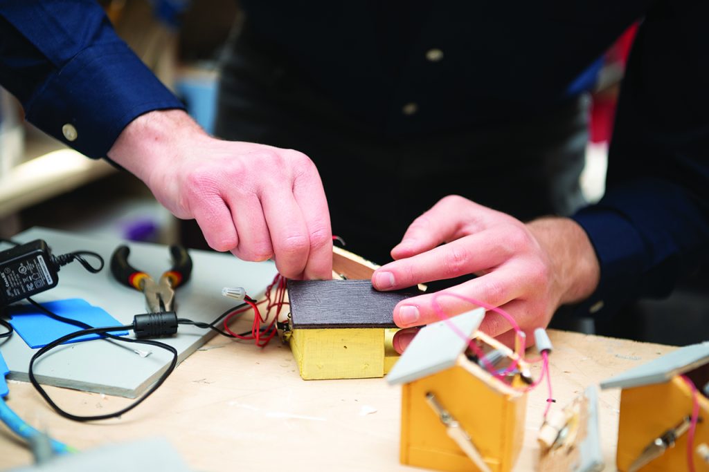 A close-up of a student’s hands connecting a red wire to a miniature wooden house. Pliers, extra wires, and partially constructed models are scattered around the work space.