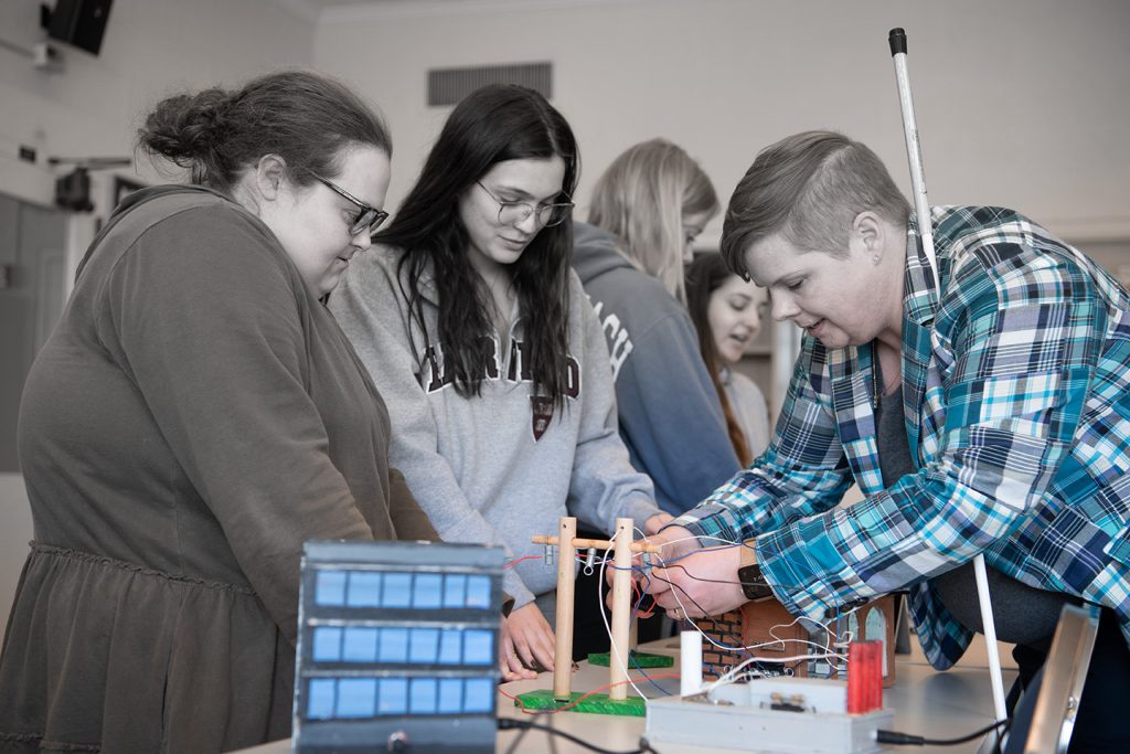 Two students observe as Dr. Natalie Shaheen connects a red wire to a miniature telephone pole within the nonvisually accessible Smart Grid for All kit.