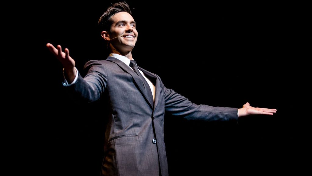 Michael Carbonaro wearing a suit on stage with his arms extended