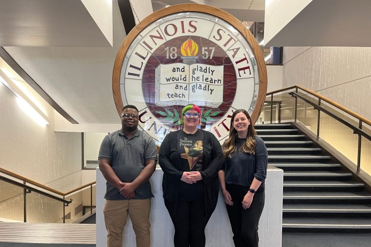 Three staff people standing in front of the stained glass ISU seal on the 2nd floor of Milner Library