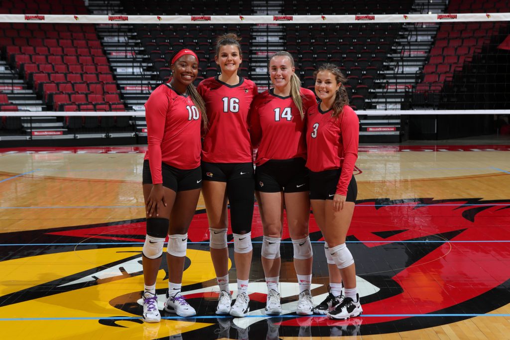 Four volleyball players wearing red uniforms pose in front of a volleyball net on the Redbird Arena court
