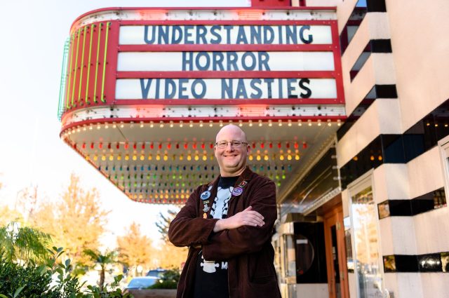 Dr. Eric Wesselmann posing in front of the Normal Theater marquee sign with text that illustrates "Understanding Horror: Video Nasties."