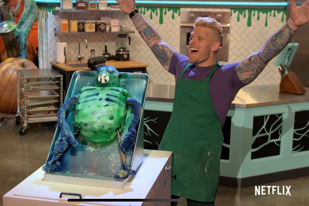 Phil Januszewski stands in front of his alien autopsy cake with his hands in the air during his presentation