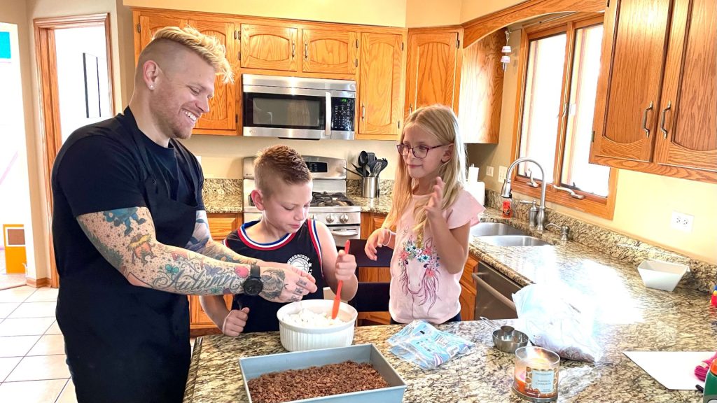 Phil and his two children stand in front of a mixing bowl and cake as they are baking