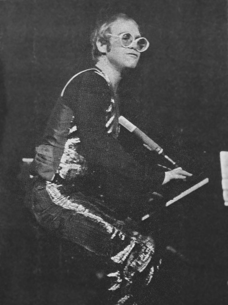 Black and white photo of Elton John standing, playing the piano