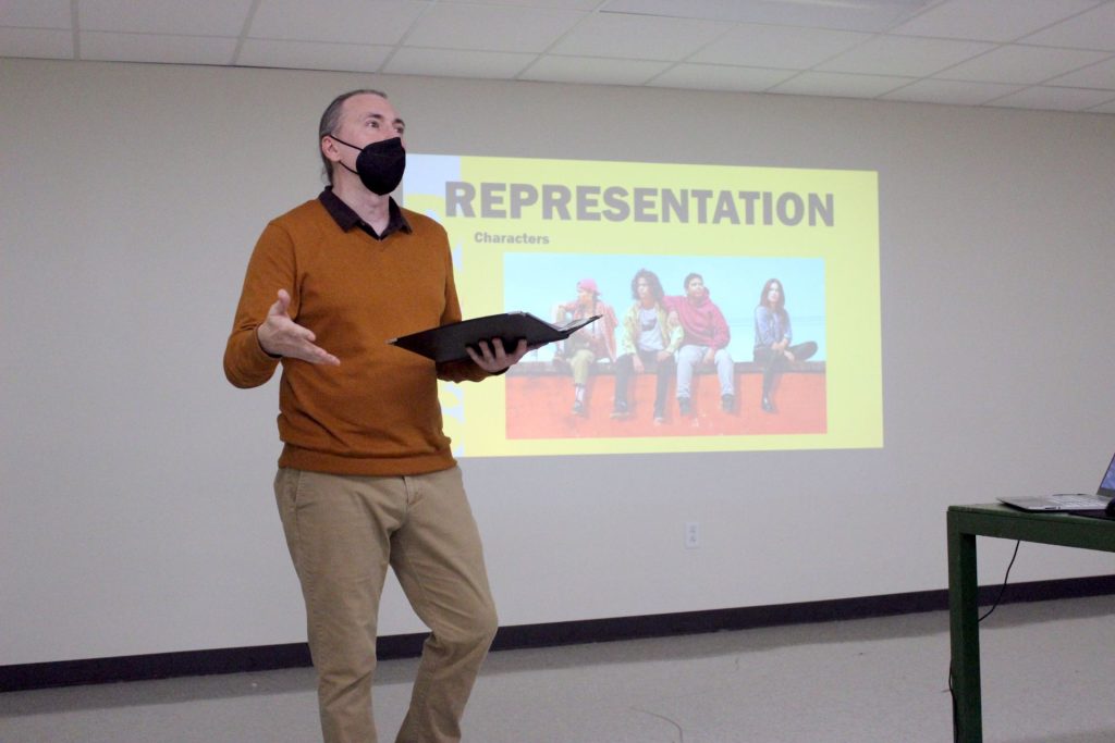 Dr. Shannon Epplett speaks in front of a projected screen with the title "Dogs Representation" and an image of the lead characters for "Reservation Dogs."