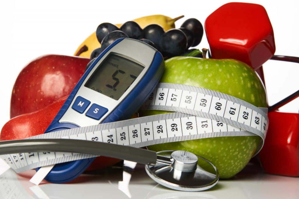 Photo of fruit, a blood glucose reader, a stethescope, and weights with a tape measure wrapped around.