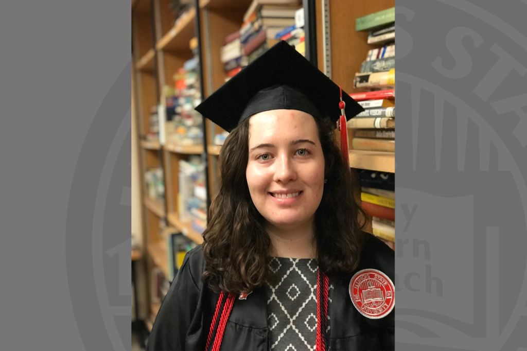 Female undergraduate student in cap and gown in front of book cases.