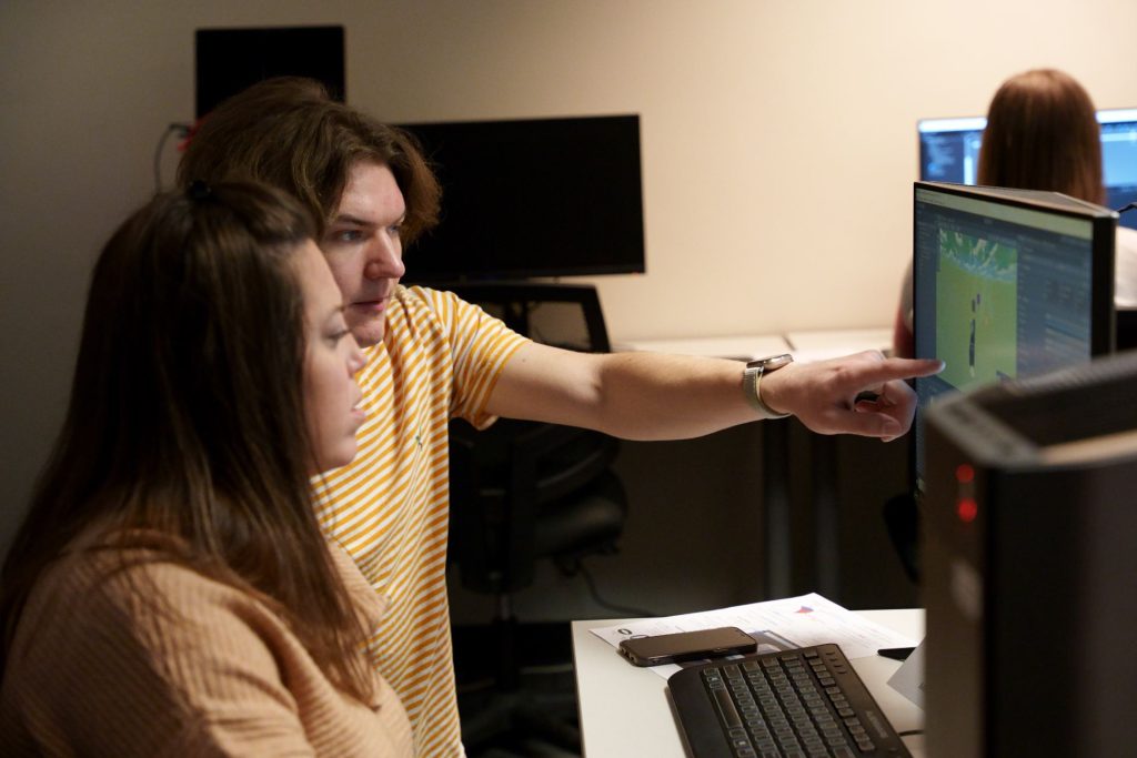 Two individuals work together on an virtual reality project that can partially be seen on a computer screen