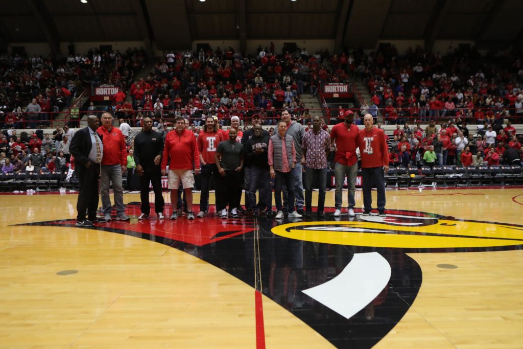 Sixteen former men's basketball players standing at center court