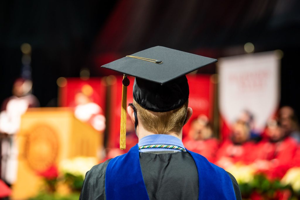 The back of a student's head, wearing a mortarboard with the commencement stage in the back