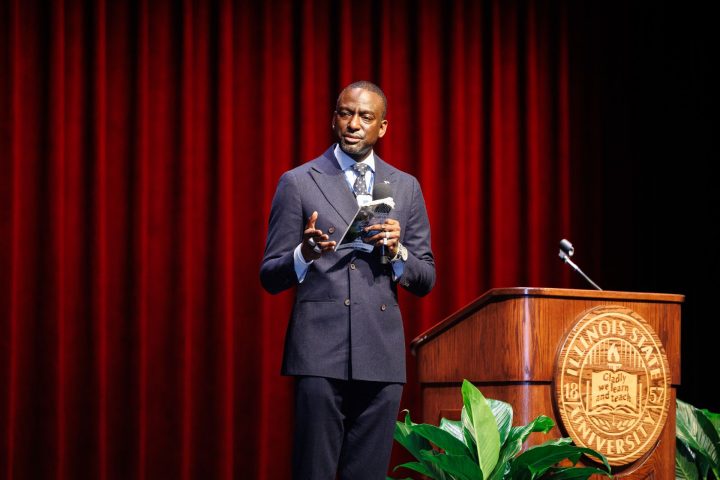 Dr. Yusef Salaam, wearing a suit and tie, speaks into a microphone while holding a book