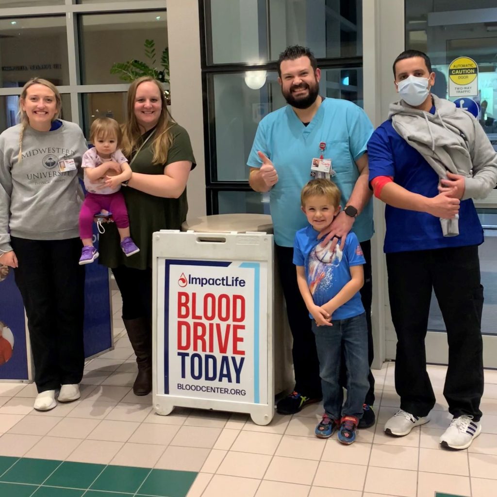 four adults and two children posing together behind a sign that reads "BLOOD DRIVE TODAY."