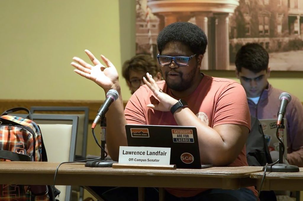 Lawrence Landfair speaking into a microphone, sitting at a table in front of a laptop computer