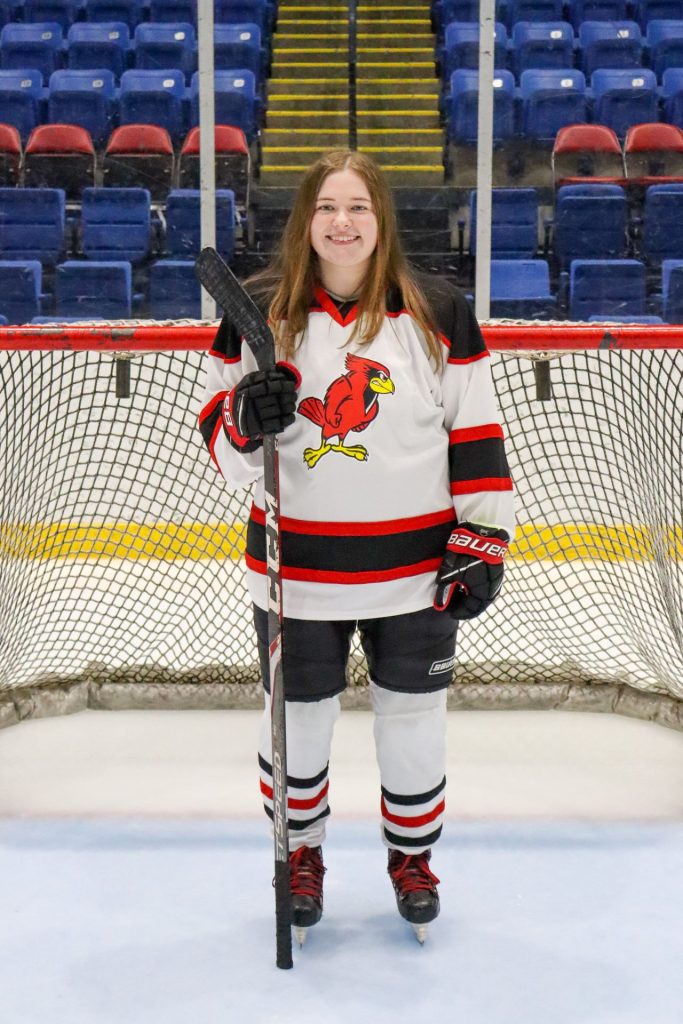 Makenna Spearman standing on ice in front of a hockey net holding a hockey stick wearing a Redbird hockey sweater