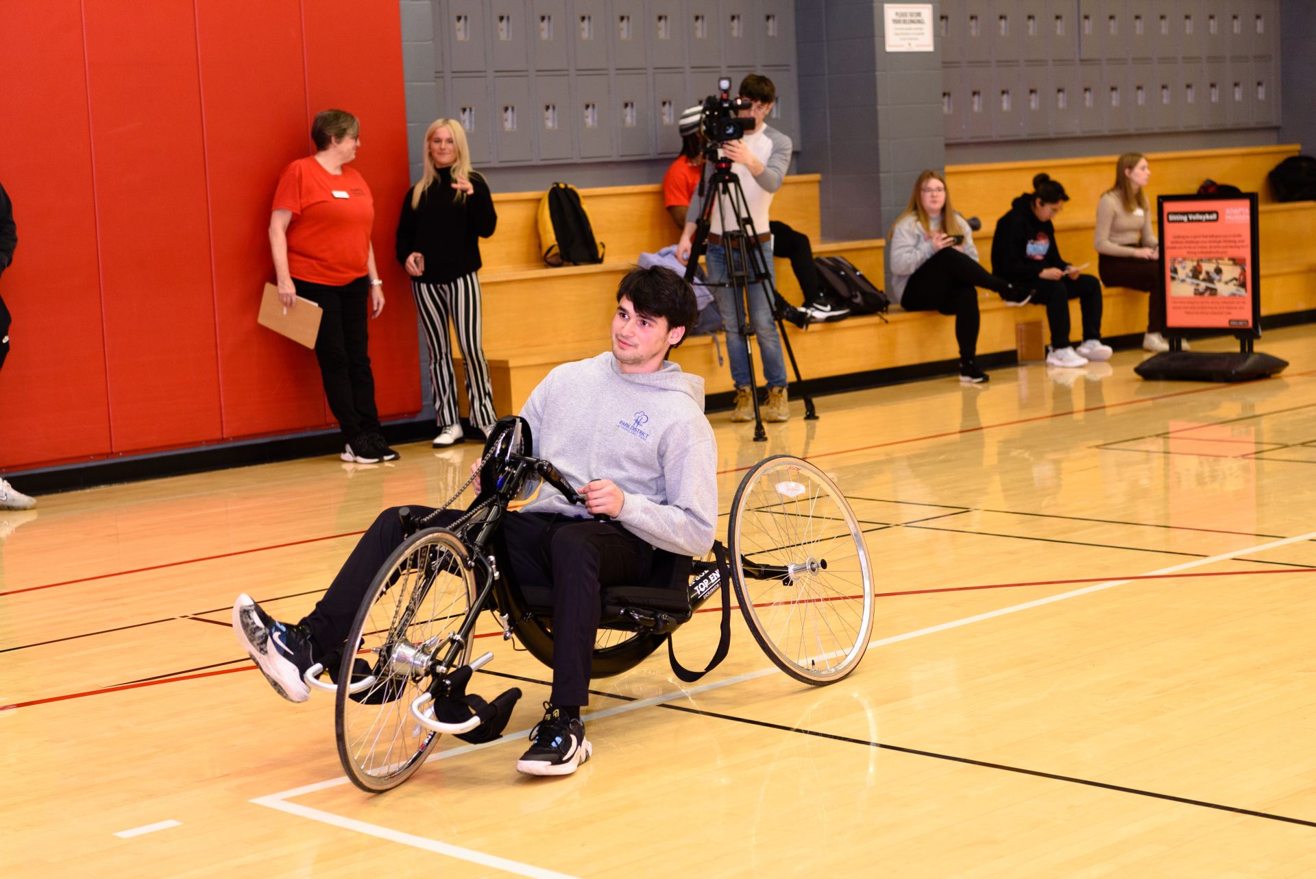 Student sitting in hand cycling chair peddles around the gym