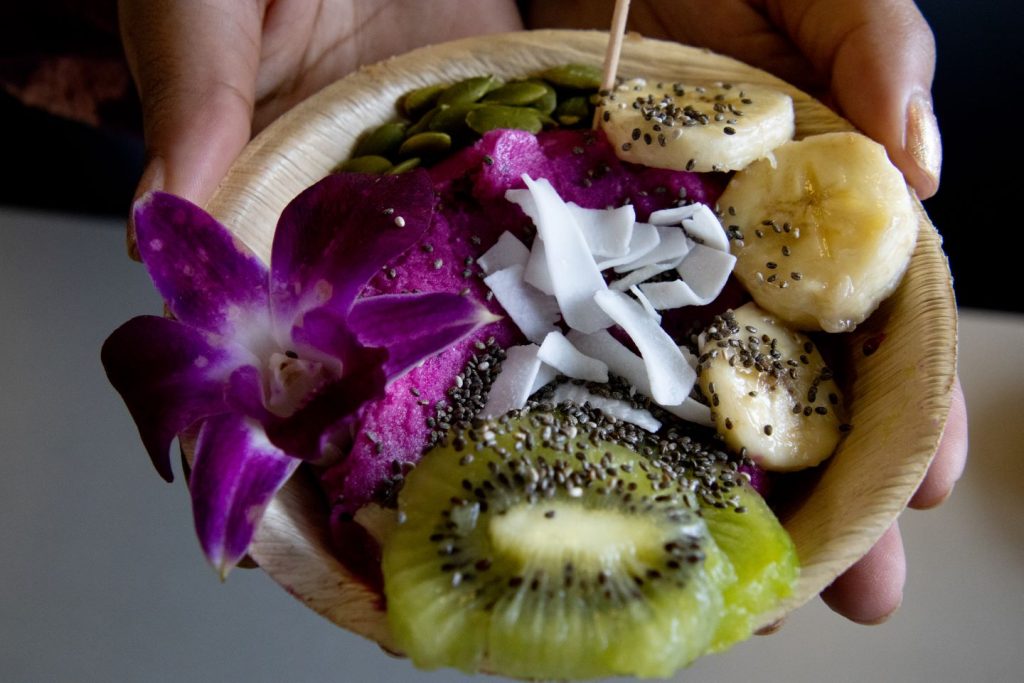 Red dragon fruit smoothie bowl topped with fresh bananas, kiwi, edible orchids, pepitas, chia seeds, and desiccated coconuts in a wooden bowl being held in someone's hands.