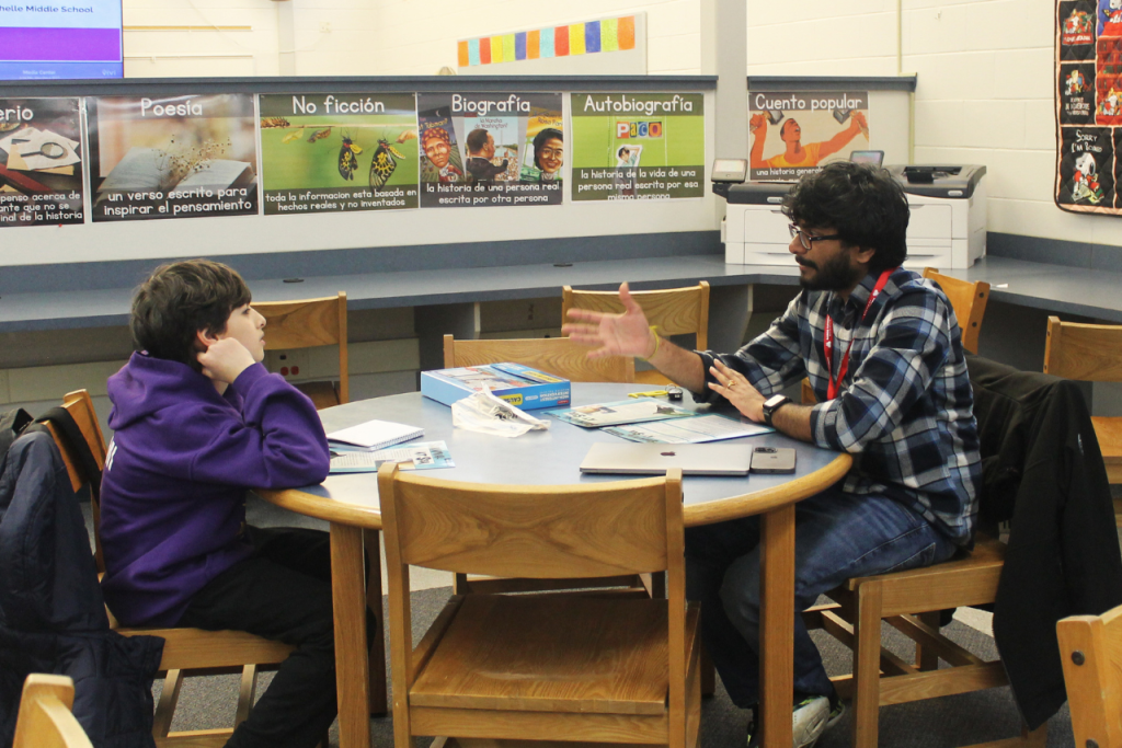 A boy and young man sit at a library table with brightly colored worksheets spread out between them. The man gestures with his hands as he explains a literary concept to the boy.