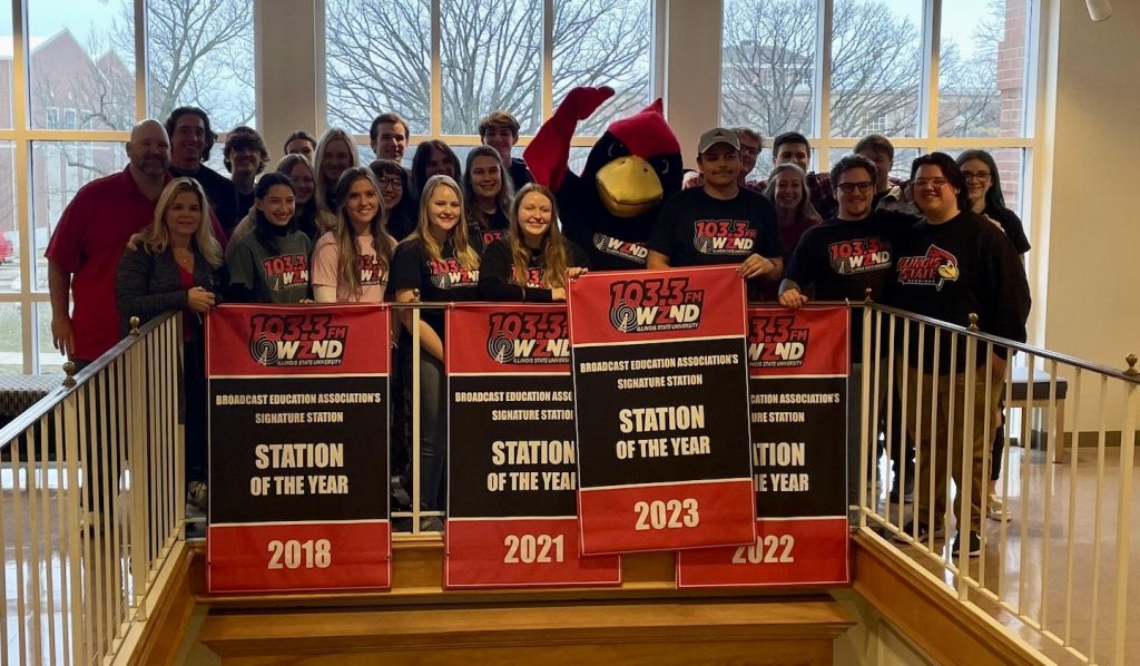 The 2023 staff of WZND Radio with Reggie Redbird and their new BEA Signature Station Banner at the second floor Atrium in Fell Hall.