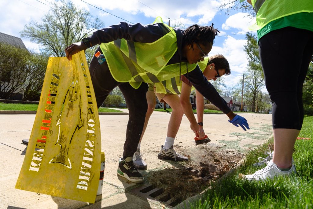A student uses a broom to clear debris from a drain, while another holds a large stencil with the outline of a fish and the words "Drains to stream, keep it clean."
