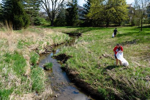 Two students use trash pickers to pick up trash and dump it in trash bags along a narrow, winding creek
