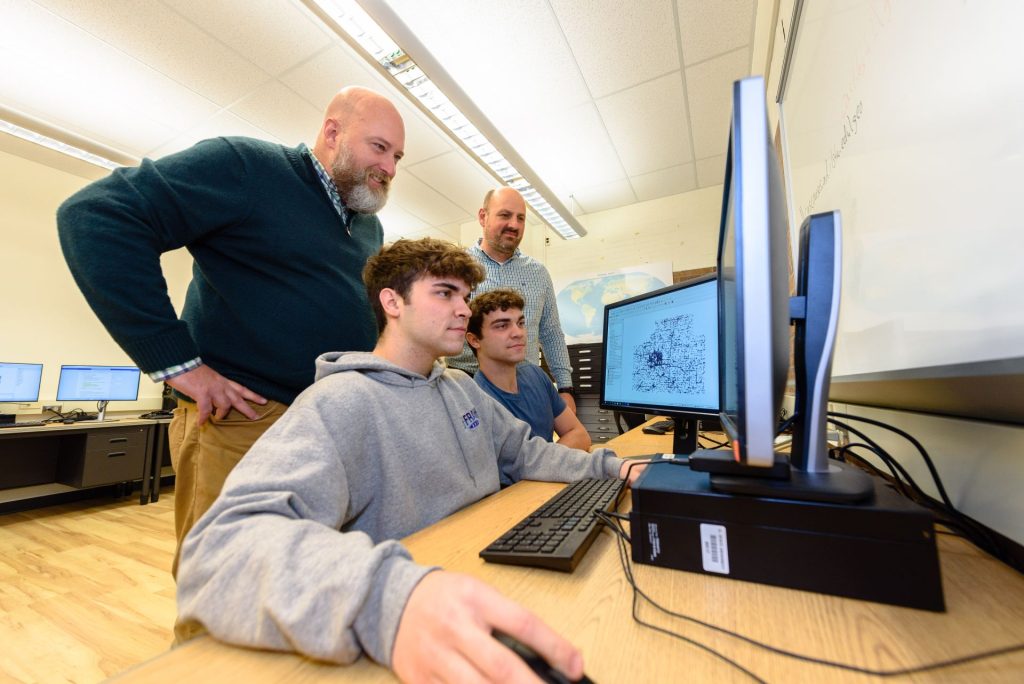 two students look at a computer screen while two professors observe standing just behind them
