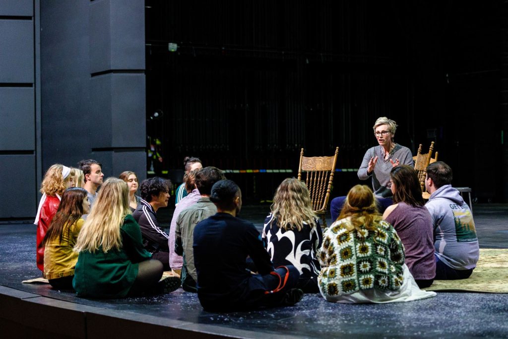 Jane Lynch, seated in a chair, talks to a group of cast and crew gathered around her, seated on the ground.
