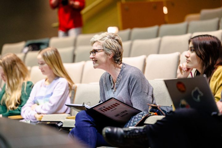 Jane Lynch smiles while sitting in the audience, holding a binder containing a script