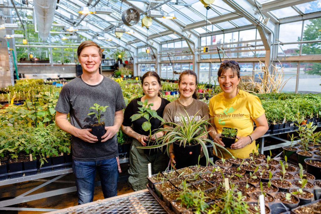 Three students and a staff member pose in a greenhouse while each holding a plant.