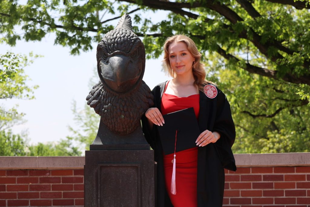 photo of Connolly in Commencement regalia standing next to the Reggie head statue on Redbird Plaza