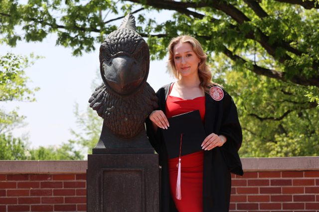 photo of Connolly in Commencement regalia standing next to the Reggie head statue on Redbird Plaza