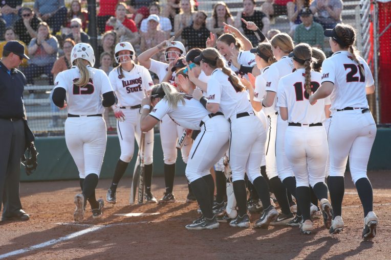 Softball players crowd around home plate to congratulate Emme Olson as she crossed home plate after a home run.