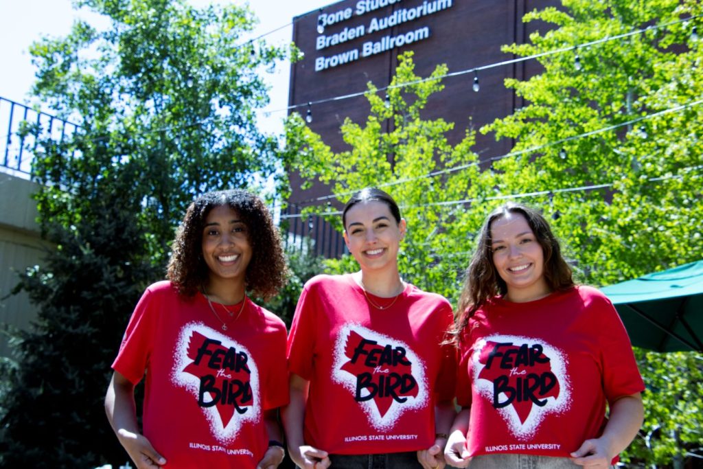 Three students model the 2023 Fear the Bird shirt in the courtyard at the Bone Student Center surrounded by trees with the Bone Student Center signage visible on the building behind them.