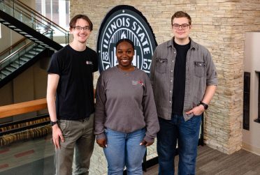 Evan Hazzard, Shira Archie, and Russell Roberts pose in front of an Illinois State University seal.