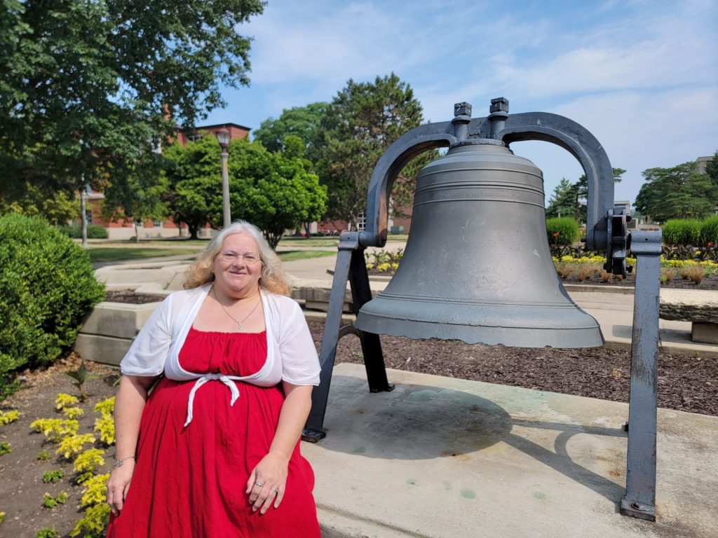 Judith Webster posing in front of the bell on the Quad.