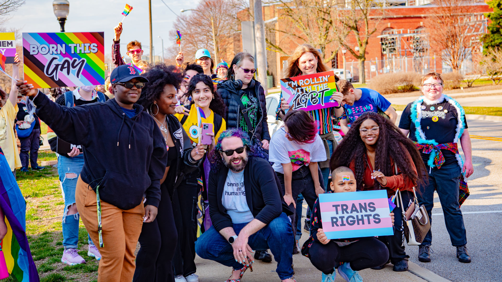 People pose together during an event holding LGBTQIA2S+ advocacy signs.