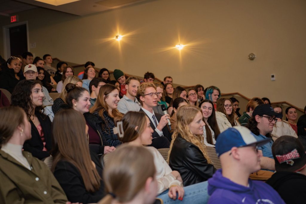 A student asks a question, speaking into a microphone, seated in an audience of students
