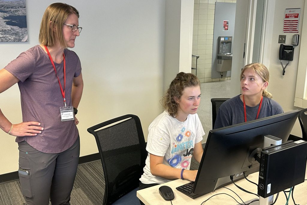 A professor offers guidance to two students sitting at computers