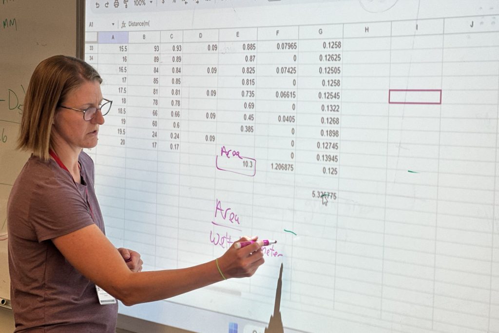 A professor writes on a whiteboard over which a spreadsheet is projected
