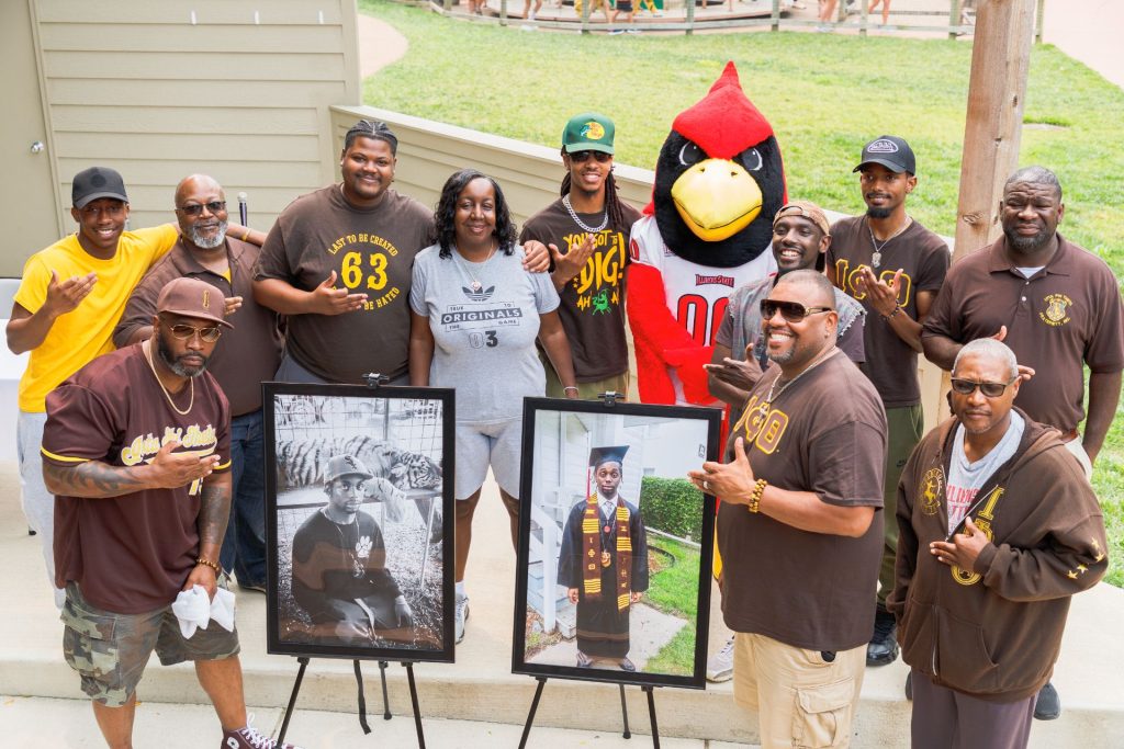 Eleven people pose with Reggie Redbird while standing by two portraits of Kristian Philpotts.