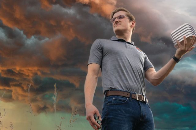 Matthew Tunberg holds internet of things weather detection device while standing in a field with a thunderstorm brewing in the background