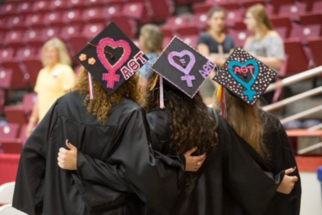 Three people standing with arms around each other, in caps and gowns, looking away from the camera with long brown hair and feminist pictures on the top of their caps.
