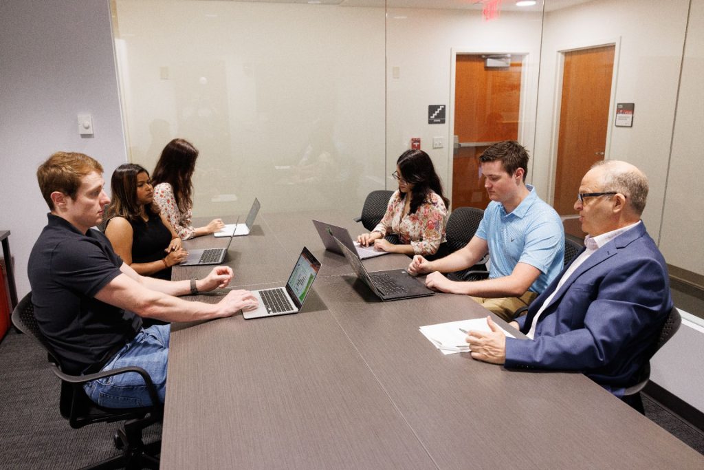 Five students and a professor sit at a conference table while discussing a marketing plan.