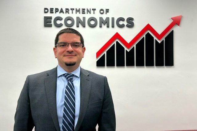 Dr. Dimitrios Nikolaou standing in front of a wall with an upward trending line and the words "Department of Economics"